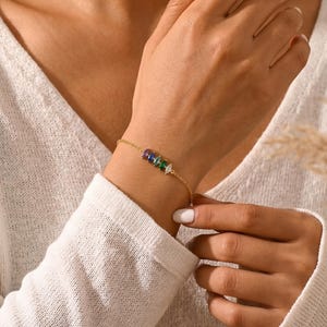 Könnte beinhalten: Ein zartes goldenes Armband mit einer Reihe farbiger Edelsteine. Das Armband zeigt eine Reihe von lila, blauen, grünen und weißen Steinen. Das Armband wird am Handgelenk getragen und hebt das Design und die Farbe des Schmucks hervor.