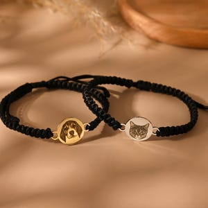 Peut inclure: Deux bracelets noirs tressés avec des pendentifs circulaires. L'un est doré avec un visage de chien, l'autre est argenté avec un visage de chat. Les bracelets sont présentés sur une surface neutre.