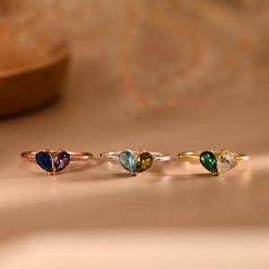 Puede incluir: Cuatro anillos con piedras preciosas en forma de corazón en varios colores: azul, morado, verde azulado, verde oliva, verde esmeralda y transparente. Los anillos tienen bandas doradas y plateadas. Los anillos se exhiben sobre una superficie neutra.