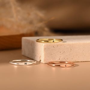 May include: Three delicate rings in gold, silver, and rose gold, each with a single initial engraved on the band. The rings are displayed on a neutral surface, showcasing their minimalist design and elegant appeal.