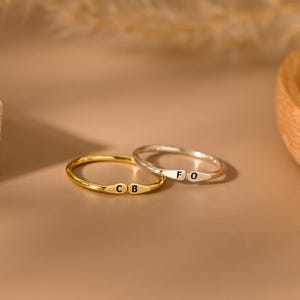 Puede incluir: Dos anillos delicados, uno dorado y otro plateado, cada uno con iniciales grabadas. El anillo dorado muestra "CB", mientras que el anillo plateado muestra "FO". Los anillos están sobre un fondo cálido y neutro, sugiriendo un tema de joyería o accesorios.