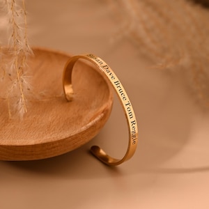 Puede incluir: Una pulsera de brazalete dorada con los nombres "Susan Dave Bruce Tom Reggie" grabados. La pulsera descansa sobre un plato de madera. El fondo es de un color suave y neutro.