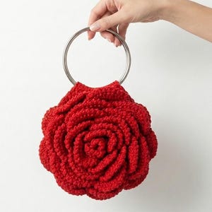 May include: A red crocheted bag shaped like a rose, held by a silver metal handle. The bag's petals are intricately detailed, and the overall design is a vibrant red. The bag is a unique accessory.