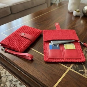 May include: Two red crochet wallets with leather accents are displayed on a wooden table. One wallet is closed with a leather strap, and the other is open, revealing card slots and a phone. Both have wrist straps.