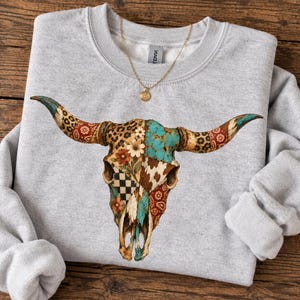 May include: A light gray crewneck sweatshirt featuring a large graphic of a cow skull. The skull is decorated with a patchwork design of leopard print, turquoise, brown cowhide, black and white checkerboard, and red floral patterns.