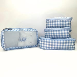 May include: A set of travel organizers in a light blue and white gingham pattern. One organizer is rectangular with a mesh front, containing a white sweater with blue embroidered initials. The other organizers are zippered, in various sizes.