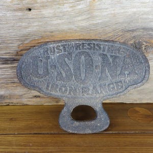 May include: A dark gray, oval-shaped cast iron stove plate featuring the embossed text RUST RESISTING U-SO-NA IRON RANGE. The object has a textured, weathered surface and a central circular cutout at the bottom, designed for handling or mounting.