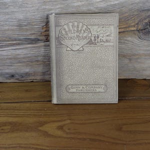 May include: A vintage book titled "The Children's Second Reader" by Ginn & Company Publishers. The book has a light gray cover with a seashell design and decorative patterns. The book is resting on a wooden surface.