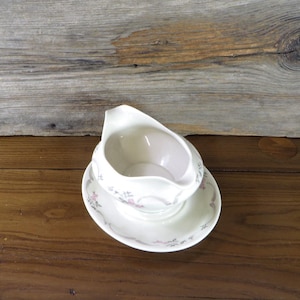 May include: A white ceramic gravy boat with a floral design on the side, sitting on a matching saucer. The boat has a rounded shape and a small spout.