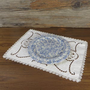 May include: A blue and white patterned plate rests on a white, lace-edged doily. The doily features a cut-out design and is placed on a wooden surface. The plate's pattern is a repeating floral design.