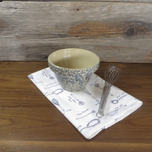 May include: A beige and blue speckled ceramic bowl sits on a white kitchen towel with blue utensil illustrations. A stainless steel whisk rests beside the bowl. The items are on a wooden surface, suggesting a kitchen or baking theme.
