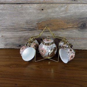 May include: A gold metal stand with a star-shaped design holds a white teapot and four matching cups. The teapot and cups are decorated with a floral pattern in shades of pink, green, and gold.