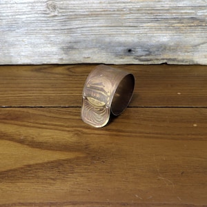 May include: A brass cuff bracelet with an etched design of a building. The bracelet is on a wooden surface.