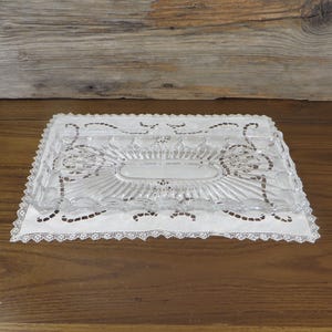 May include: A clear, rectangular glass butter dish with a decorative, ridged design, resting on a white lace doily. The doily has a scalloped edge and is placed on a wooden surface.
