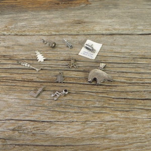 May include: A collection of silver charm pendants, including a bear, a tree, a musical note, a fish, a building, and a bell. The charms are scattered on a wooden surface.