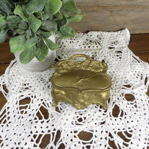 May include: A small, antique-style, gold-colored jewelry box with floral detailing and a handle. It sits on a white, open-weave lace doily, with a small potted plant in the background. The setting is on a wooden surface.