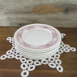 May include: A stack of six white ceramic plates with a decorative maroon floral border. The plates rest on a white, openwork lace doily. The background features a wooden surface and a weathered wood backdrop.