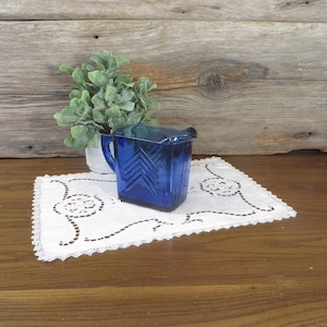 May include: A vintage cobalt blue glass pitcher with a handle and a spout, set on a white lace doily. The pitcher has a geometric design and is next to a small potted plant. The background is a wooden wall.