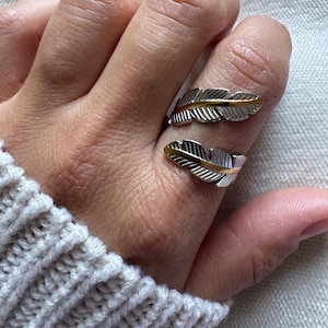 Op de afbeelding: Een open ring met twee zilverkleurige veermotieven. Elke veer heeft een goudkleurige centrale nerf over de gehele lengte, met gedetailleerde gegraveerde lijnen die uitstralen naar de randen van de metalen bladeren op de vinger.