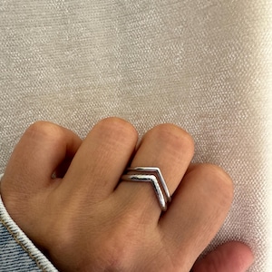 Pode incluir: Um conjunto de dois anéis prateados em forma de chevron usados num dedo. O anel superior apresenta um acabamento liso e polido, enquanto o anel inferior tem uma superfície texturizada e martelada. Ambos formam um design em forma de V.