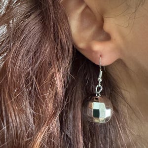 Può includere: Orecchini a sfera da discoteca argentati con superficie riflettente. Gli orecchini sono appesi a un gancio d'argento. Gli orecchini sono rotondi e hanno un design sfaccettato. Indossati da una persona con i capelli castani.