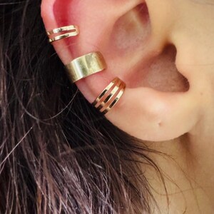 Peut inclure: Trois bagues d'oreille dorées portées sur le cartilage externe. La bague supérieure présente deux fines bandes, la bague centrale est une large bande pleine, et la bague inférieure se compose de trois fines bandes parallèles, finition métallique.