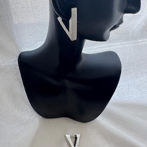 May include: A pair of silver-toned triangular hoop earrings with a hinged closure. One earring is displayed on a black mannequin bust, while the other is positioned separately below, showcasing the geometric shape and metallic finish of the jewelry.