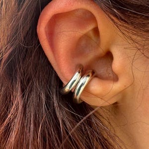 Op de afbeelding: Zilveren oormanchet sieraden. De oormanchet heeft een dubbelbandontwerp dat om het oor gewikkeld is. De sieraden zijn glanzend en reflecterend, met een glad, afgerond oppervlak. De oormanchet wordt op het oor gedragen, met bruin haar op de achtergrond.