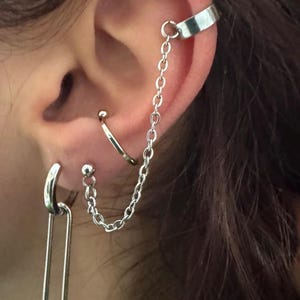 Puede incluir: Juego de joyas para la oreja de plata con un brazalete, cadena, aro y pendientes de botón. El brazalete se envuelve alrededor de la parte superior de la oreja, conectado por una cadena a un botón. También se ve un aro y un pendiente de botón.