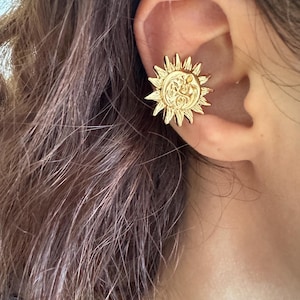 May include: A gold-toned ear cuff featuring a circular sun design with a central face and radiating pointed rays. The metal piece is positioned on the outer cartilage of the ear, showcasing a polished finish and detailed embossed features.