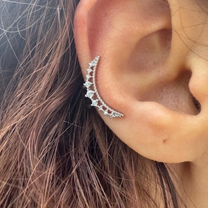 May include: A silver-toned ear cuff shaped like a crescent moon, featuring a delicate filigree metal design and a series of small, clear, round-cut stones set along the outer edge, worn along the cartilage of the ear.