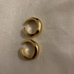 K&ouml;nnte beinhalten: Zwei goldfarbene Ohrklemmen mit glatter, polierter Oberfl&auml;che und C-f&ouml;rmigem Design. Jedes St&uuml;ck besteht aus einem dicken, abgerundeten Band mit einer &Ouml;ffnung, wodurch eine durchgehende, leicht unregelm&auml;&szlig;ige Kreisform entsteht.