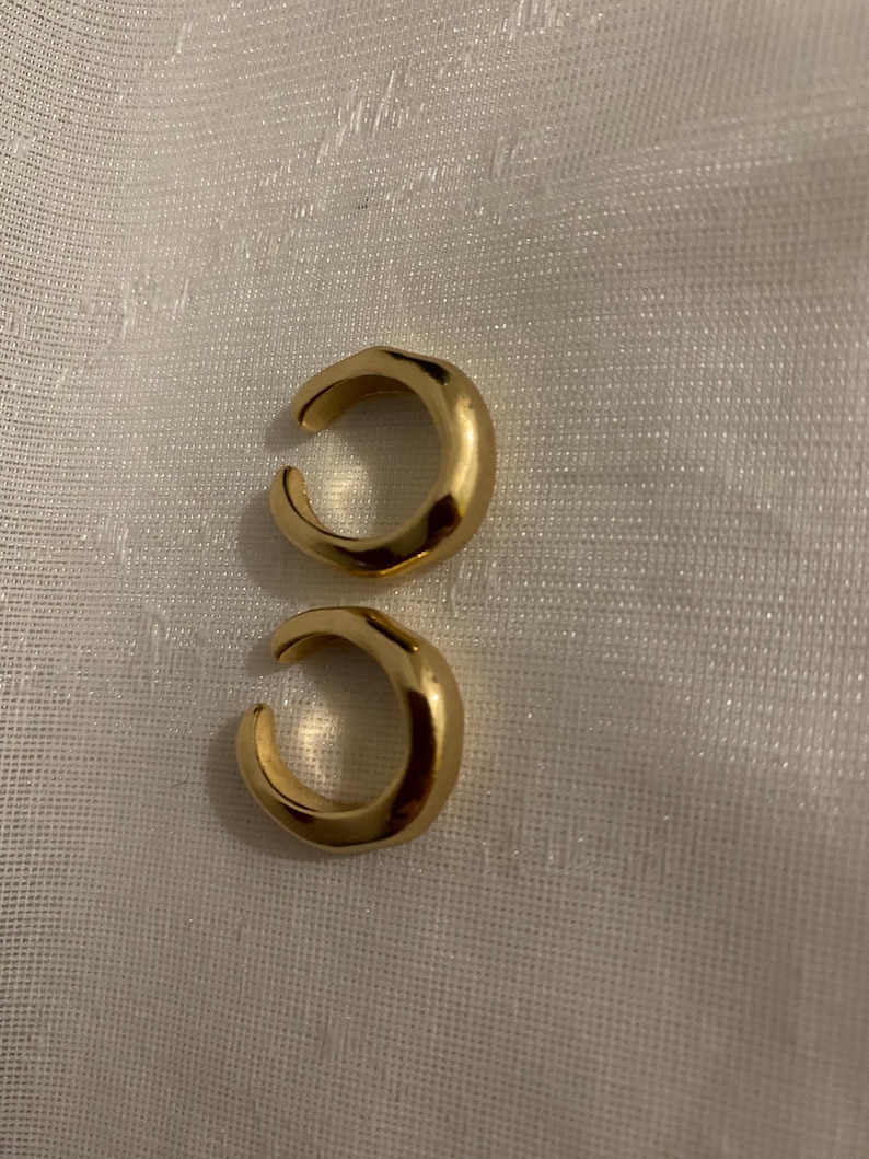 K&ouml;nnte beinhalten: Zwei goldfarbene Ohrklemmen mit glatter, polierter Oberfl&auml;che und C-f&ouml;rmigem Design. Jedes St&uuml;ck besteht aus einem dicken, abgerundeten Band mit einer &Ouml;ffnung, wodurch eine durchgehende, leicht unregelm&auml;&szlig;ige Kreisform entsteht.