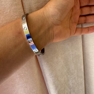 Op de afbeelding: Een zilverkleurige metalen armband met een gladde, gepolijste afwerking. De band is voorzien van gelijkmatig verdeelde, ronde zettingen met heldere, fonkelende stenen en afgewisseld met klaverbladvormige uitsparingen op het oppervlak.