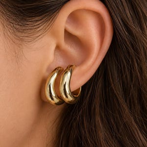 K&ouml;nnte beinhalten: Ein Paar goldfarbene Ohrklemmen mit einer glatten, polierten und abgerundeten Oberfl&auml;che. Der Schmuck zeichnet sich durch ein dickes, geschwungenes Design aus, das sicher am &auml;u&szlig;eren Rand des Ohrknorpels sitzt und einen geschichteten Look mit zwei B&auml;ndern erzeugt.
