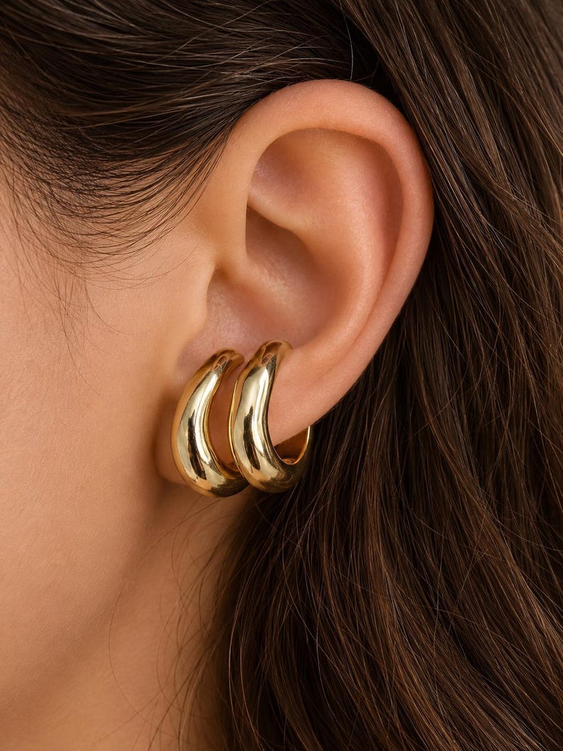 K&ouml;nnte beinhalten: Ein Paar goldfarbene Ohrklemmen mit einer glatten, polierten und abgerundeten Oberfl&auml;che. Der Schmuck zeichnet sich durch ein dickes, geschwungenes Design aus, das sicher am &auml;u&szlig;eren Rand des Ohrknorpels sitzt und einen geschichteten Look mit zwei B&auml;ndern erzeugt.