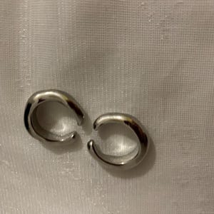 K&ouml;nnte beinhalten: Ein Paar silberfarbene Ohrklemmen mit glatter, polierter Oberfl&auml;che und offenem, kreisf&ouml;rmigem Design. Jedes St&uuml;ck besteht aus einem dicken, abgerundeten Band, das sich zu einer C-Form biegt und ein schlichtes, reflektierendes Aussehen erzeugt.