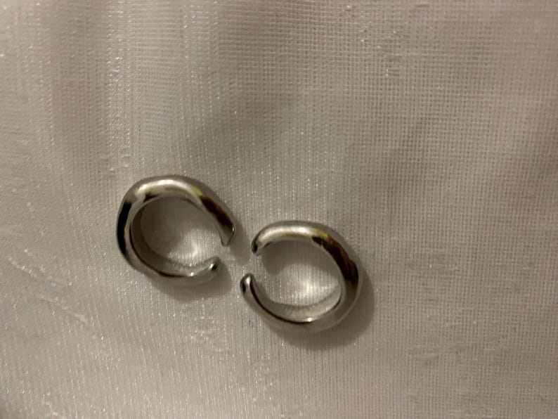 K&ouml;nnte beinhalten: Ein Paar silberfarbene Ohrklemmen mit glatter, polierter Oberfl&auml;che und offenem, kreisf&ouml;rmigem Design. Jedes St&uuml;ck besteht aus einem dicken, abgerundeten Band, das sich zu einer C-Form biegt und ein schlichtes, reflektierendes Aussehen erzeugt.