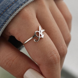 Può includere: Un anello color argento caratterizzato da una prominente lettera A maiuscola stilizzata come elemento centrale del design. La fascia è sottile e liscia, collegata alla base della lettera. Il metallo ha una finitura lucida e riflettente.