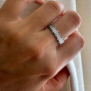 May include: A silver-toned ring featuring a series of vertical, rectangular bars arranged in a continuous, textured band. The metal surface has a polished finish, creating a repeating pattern of raised segments that encircle the finger.
