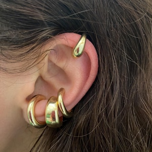 Puede incluir: Primer plano de una oreja adornada con pendientes dorados. La oreja presenta un pendiente de oreja en forma de lágrima y tres pendientes de aro gruesos y redondeados. La joyería tiene un acabado brillante y pulido, que refleja la luz.