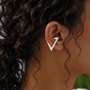 May include: A silver-toned ear cuff featuring a geometric triangle shape. The open-frame design wraps around the outer edge of the ear cartilage, creating a sharp, angular silhouette that rests securely against the side of the ear.