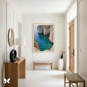 May include: A framed photograph of a turquoise blue cove with swimmers between towering cliffs hangs on a white wall. The photograph is in a light wood frame. The room has a wooden console table with a lamp and a decorative sculpture, a bench, and a door.