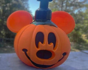 May include: An orange soap dispenser shaped like a pumpkin with round ears and a black pump top. The pumpkin features a carved face with black recessed eyes, a nose, and a wide smiling mouth. The design resembles a classic cartoon character.