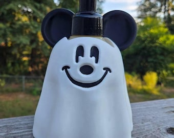 May include: A white soap dispenser shaped like a ghost with a black pump mechanism. The ghost features a black cutout design for the eyes, nose, and a wide smiling mouth, along with two prominent black circular ears extending from the sides.