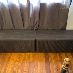 May include: Two dark gray wooden storage benches with a textured finish. The benches are rectangular with a flat top and a ribbed design on the front. They sit on a hardwood floor in front of a brown curtain.