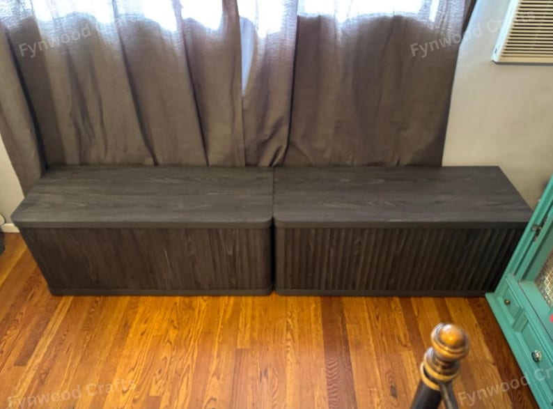 May include: Two dark gray wooden storage benches with a textured finish. The benches are rectangular with a flat top and a ribbed design on the front. They sit on a hardwood floor in front of a brown curtain.