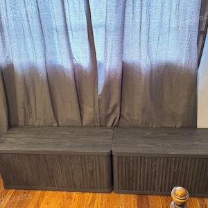 May include: Two dark gray wooden storage benches with a vertical grooved design on the front panels. The benches have flat tops and are positioned in front of a gray curtain. The benches are on a hardwood floor.
