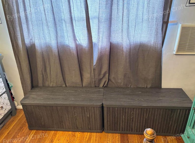May include: Two dark gray wooden storage benches with a vertical grooved design on the front panels. The benches have flat tops and are positioned in front of a gray curtain. The benches are on a hardwood floor.