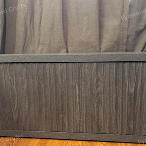 May include: A large, rectangular wooden storage chest with a dark gray wood grain finish. The chest has a flat top and a base, and is set against a neutral-colored curtain backdrop. The chest is designed for storage.